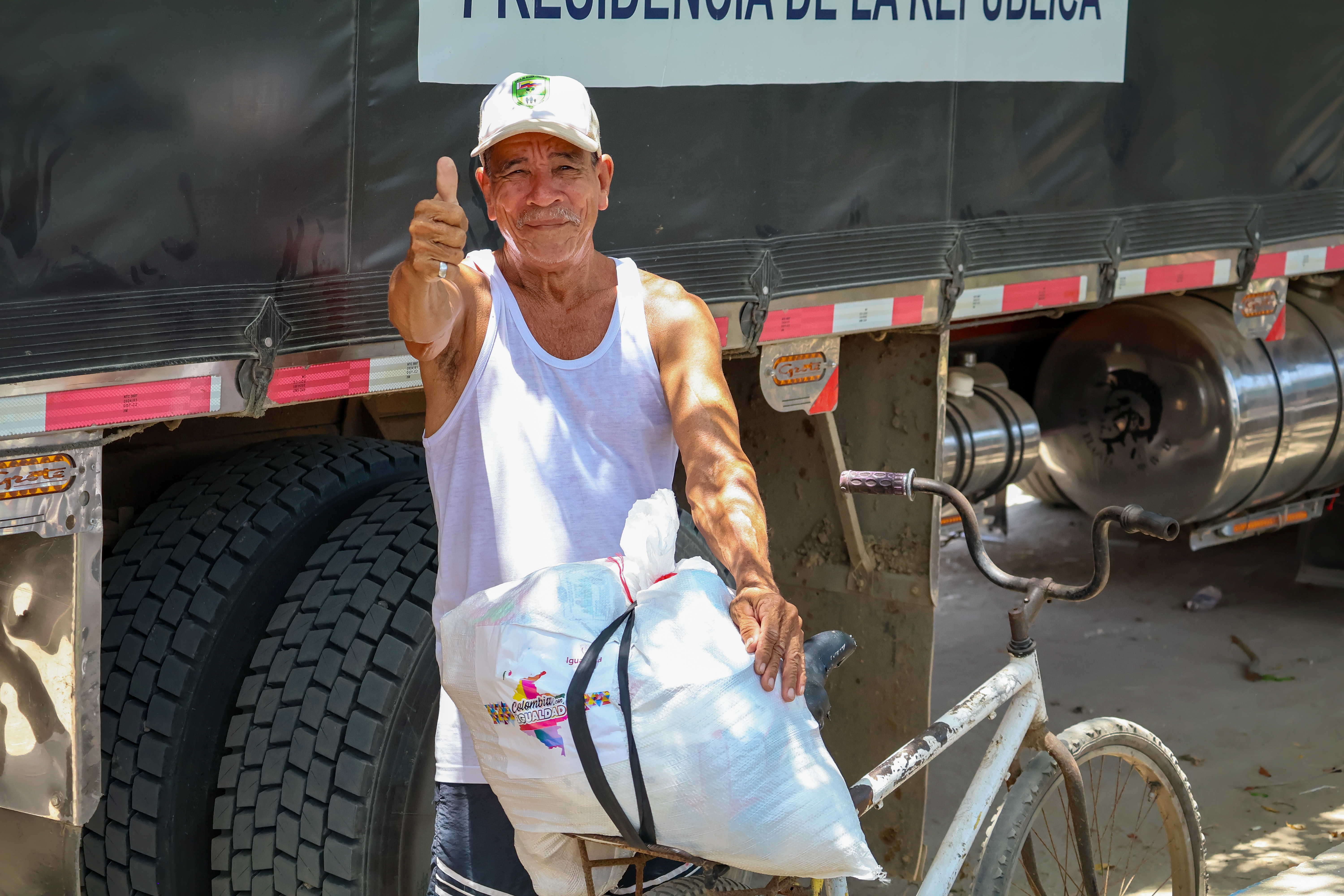 Hombre sosteniendo con su mano izquierda un costal de Colombia con Igualdad amarrado a su bicicleta y levanta su pulgar de la mano derecha reafirmando la actividad
