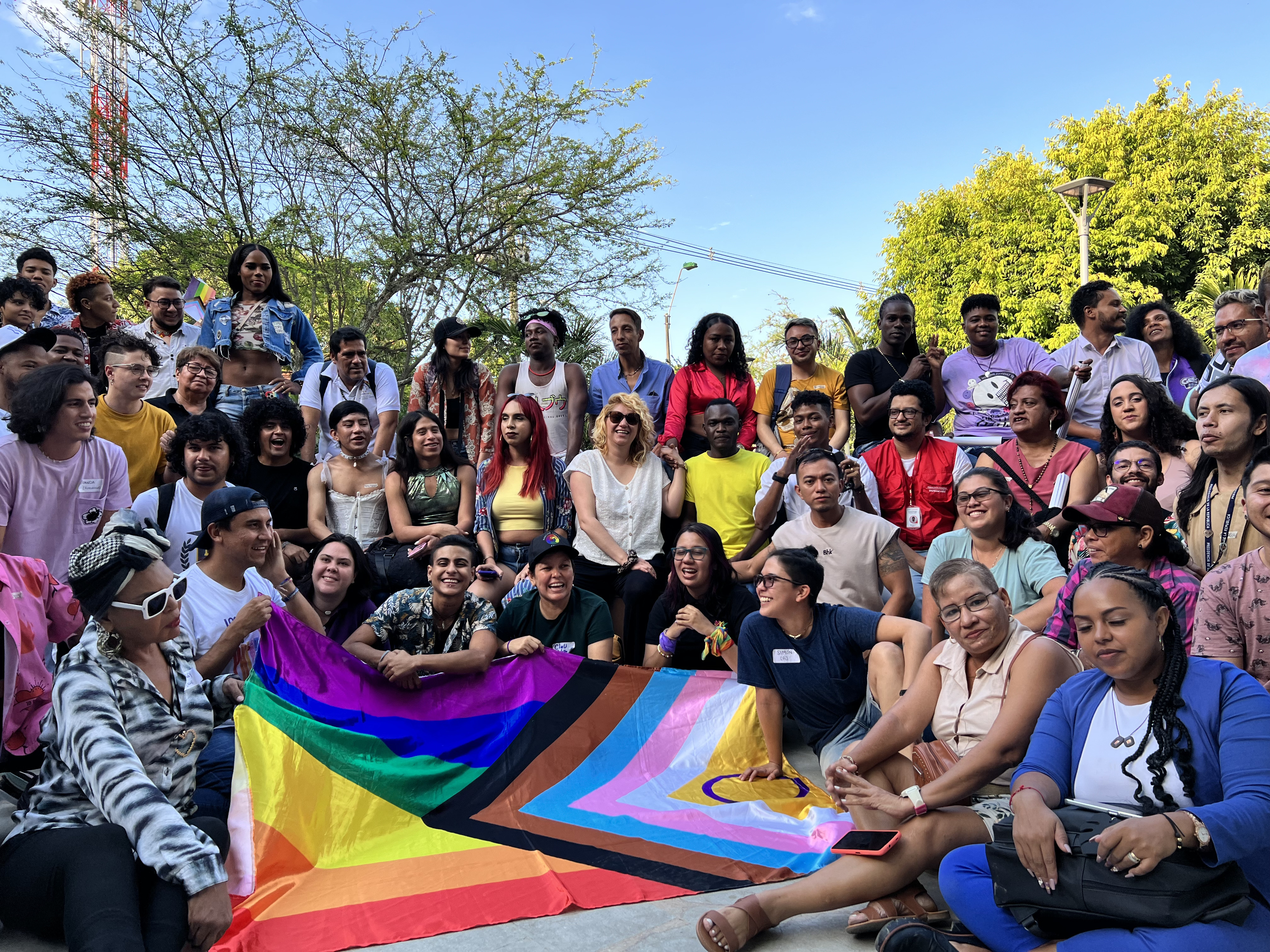 Personas con orientación sexual e identidad de género diversa posan alegres  en un espacio abierto alrededor de la bandera progress LGBTIQ+