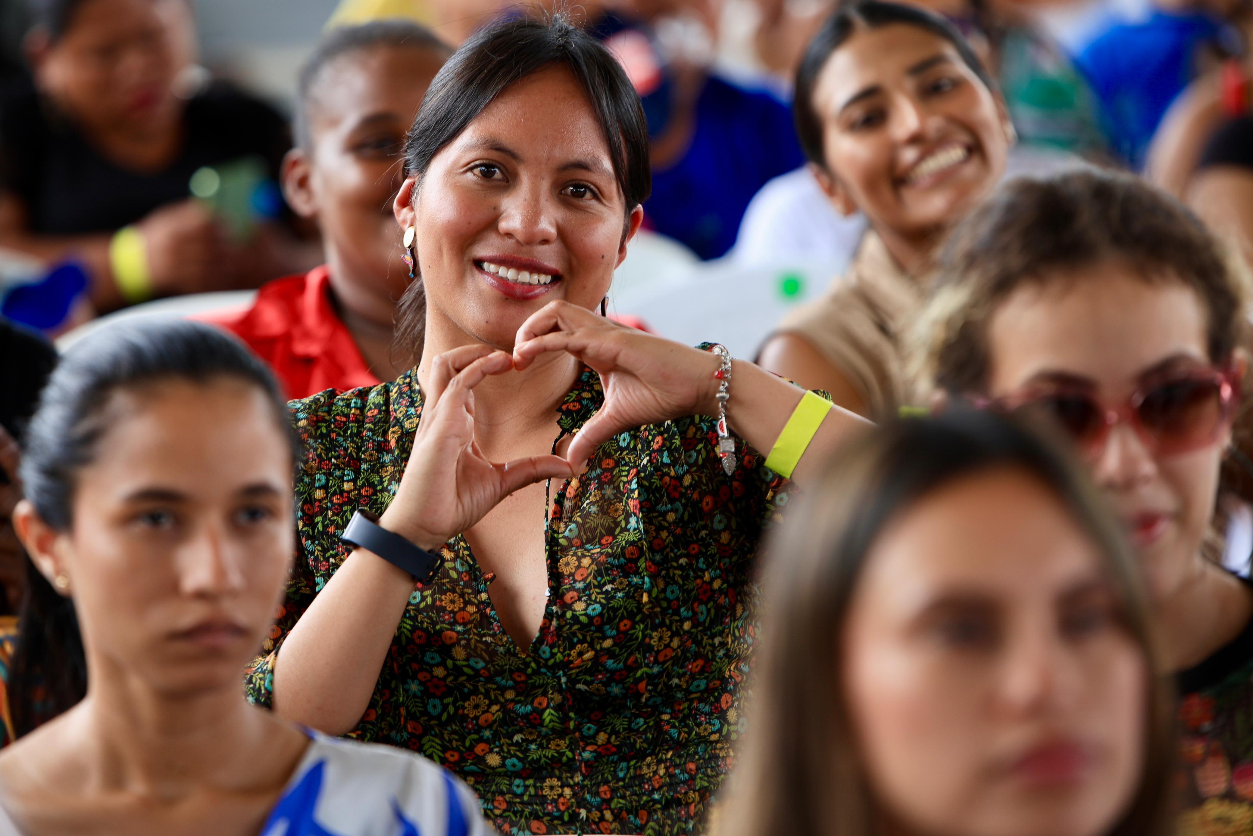 En medio de personas desenfocadas que asisten a uno de los Encuentros con el Pueblo se aprecia una mujer sonriente que mira a la cámara y con sus manos hace un corazón.