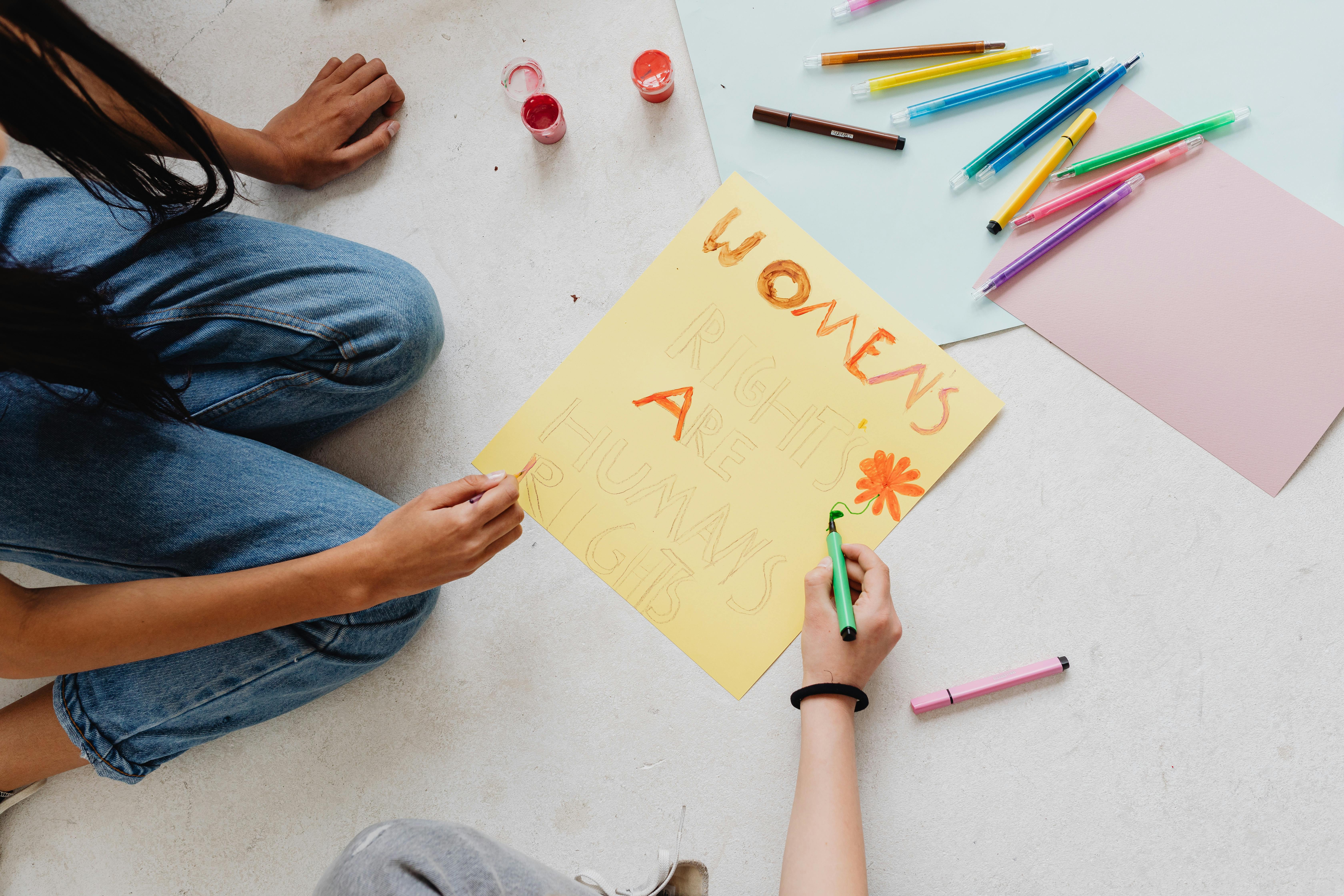 Brazos de mujeres con lápices y marcadores de color pintando un pequeño cartel sobre el piso en el que se lee la palabra women escrita con trazos manuales de varios colores