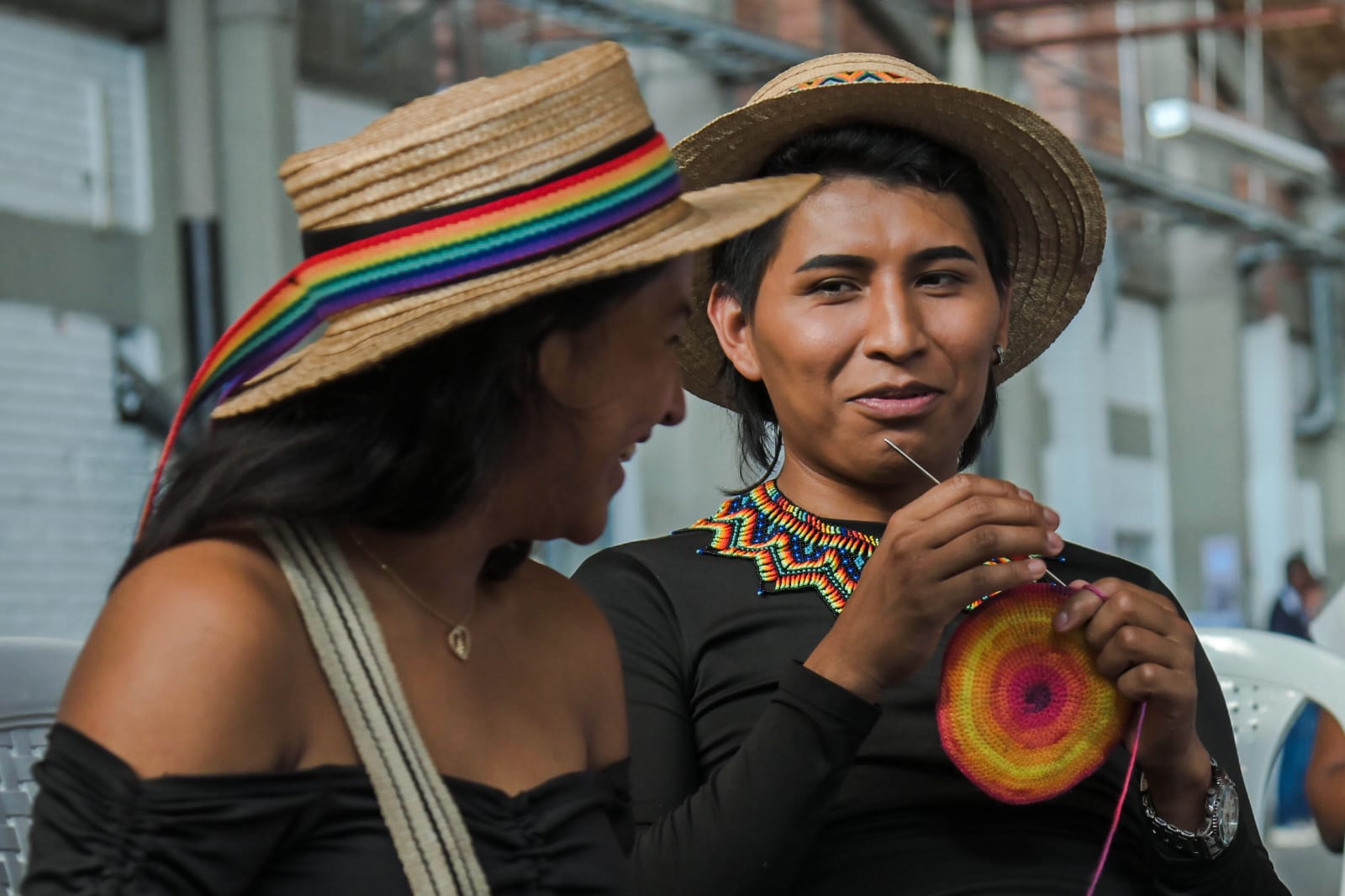 Una par de mujeres jóvenes indígeas del Cauca departen alegremente mientras una se ellas sonríe y muestra a la camara el tejido que elabora con una aguja.