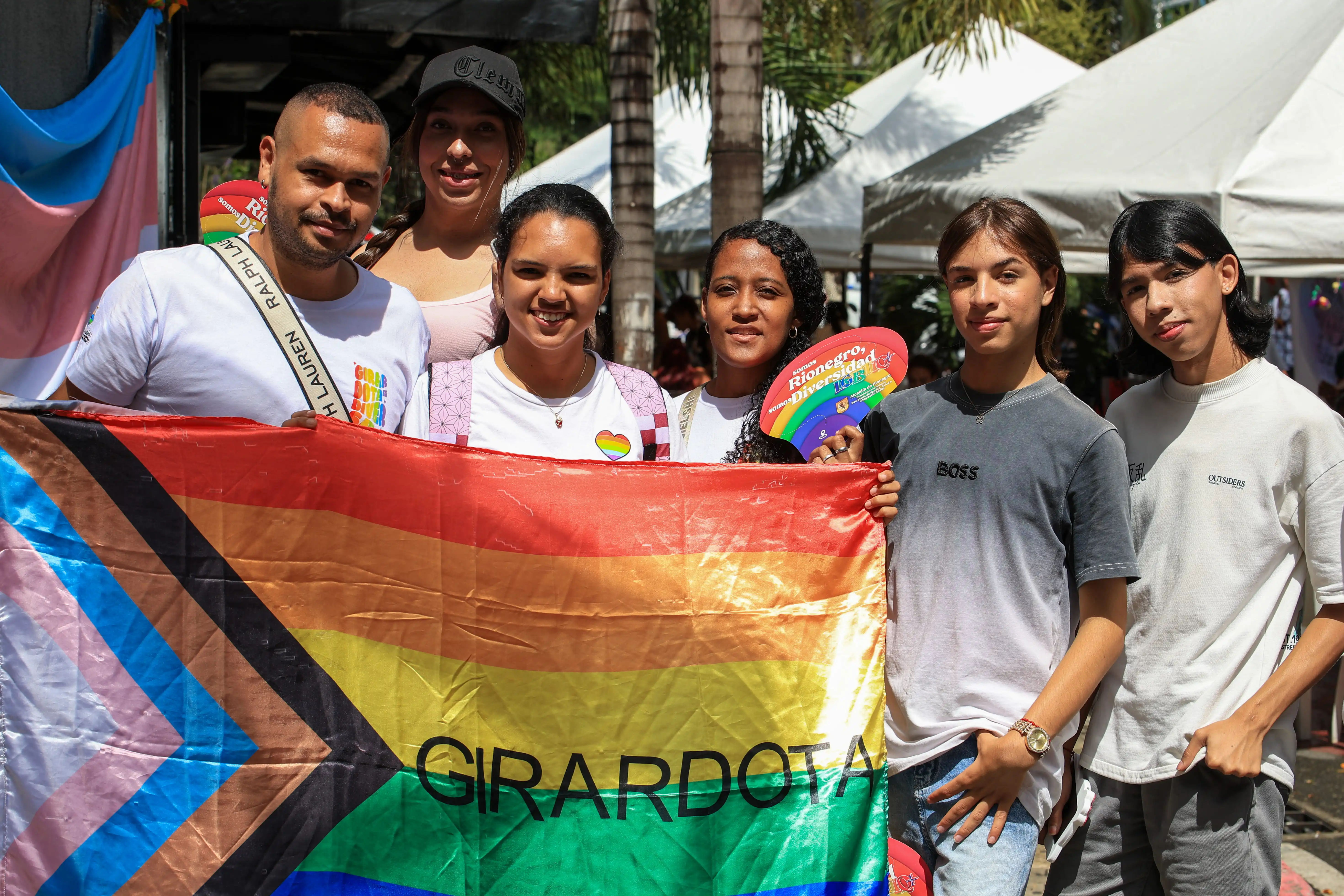 Grupo de jóvenes en una calle soleada de Medellín posa de pie detrás de una bandera LGBTIQ+ con la leyenda Girardota