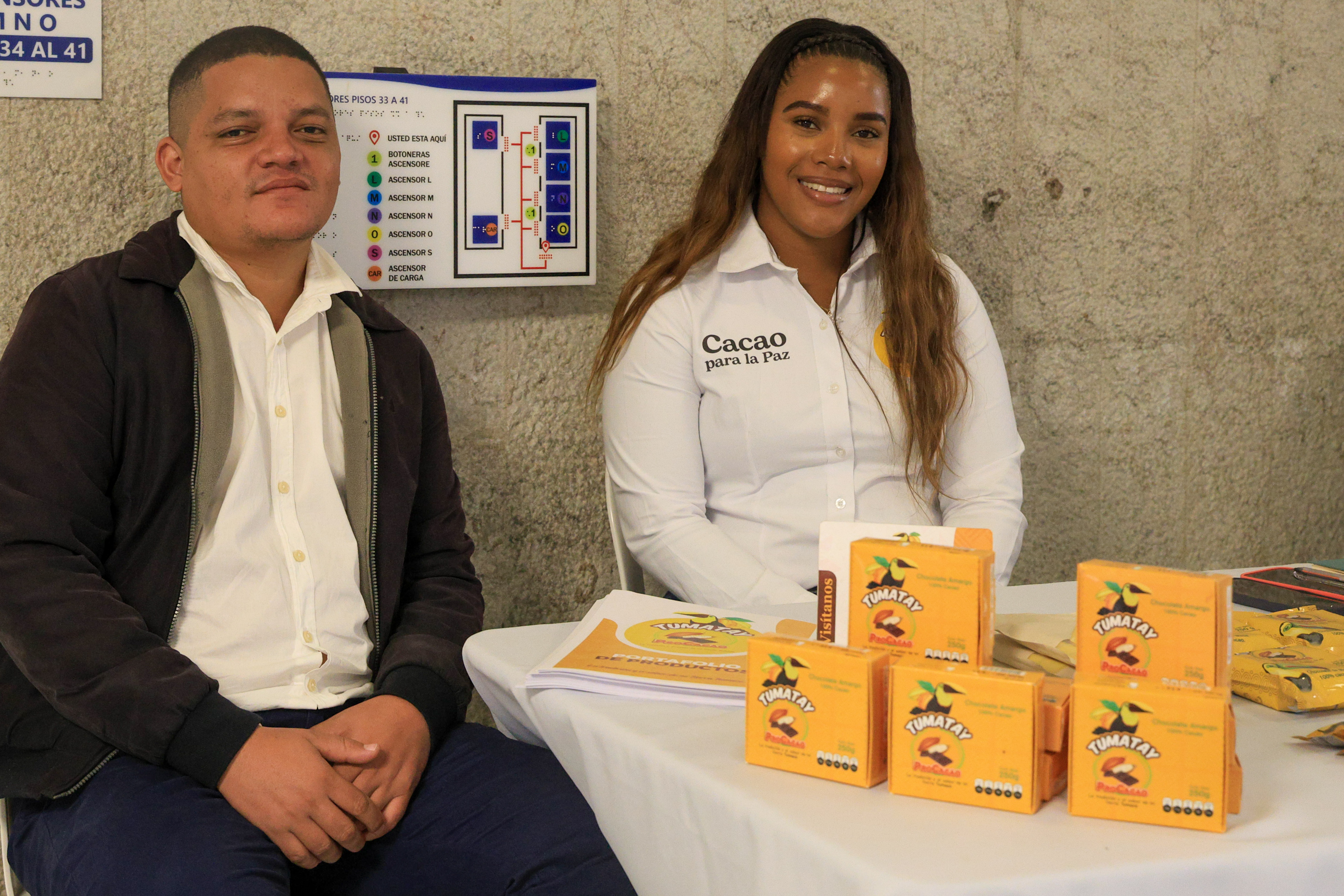 Una pareja de productores de cacao posa sentada junto a una mesa donde exhiben sus productos en medio de la feria de emprendimientos de miniguladad