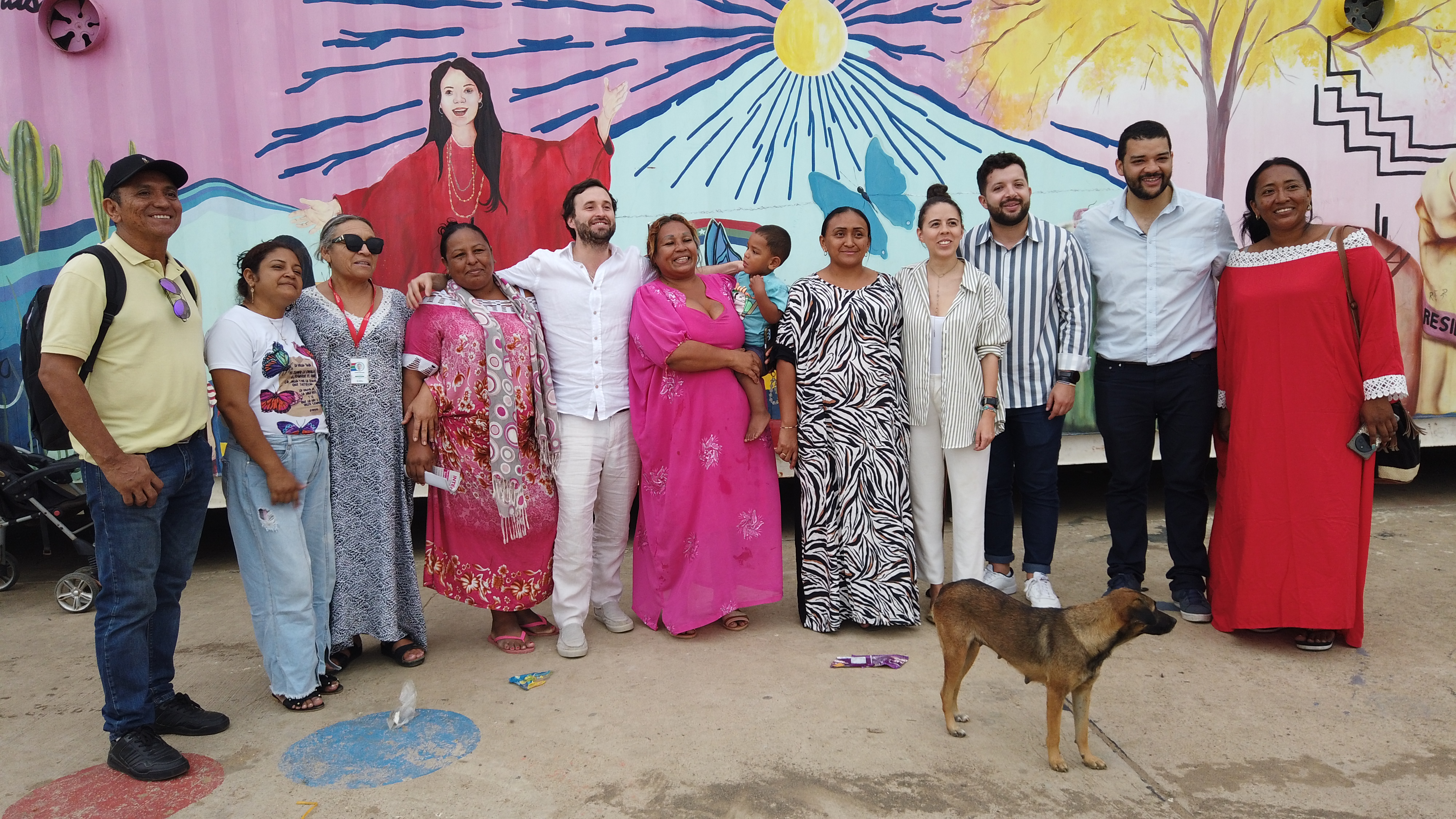 El viceministro Pablo Pardp, acompañado de su equipo de trabajo se abraza con represetantes de la comunidad migrante del antiguo sector de La Pista, en Maicao, hoy Barrio de Paz. Posan de pie por delante de una pintura de la Guajia plasmada en un contenedor de carga