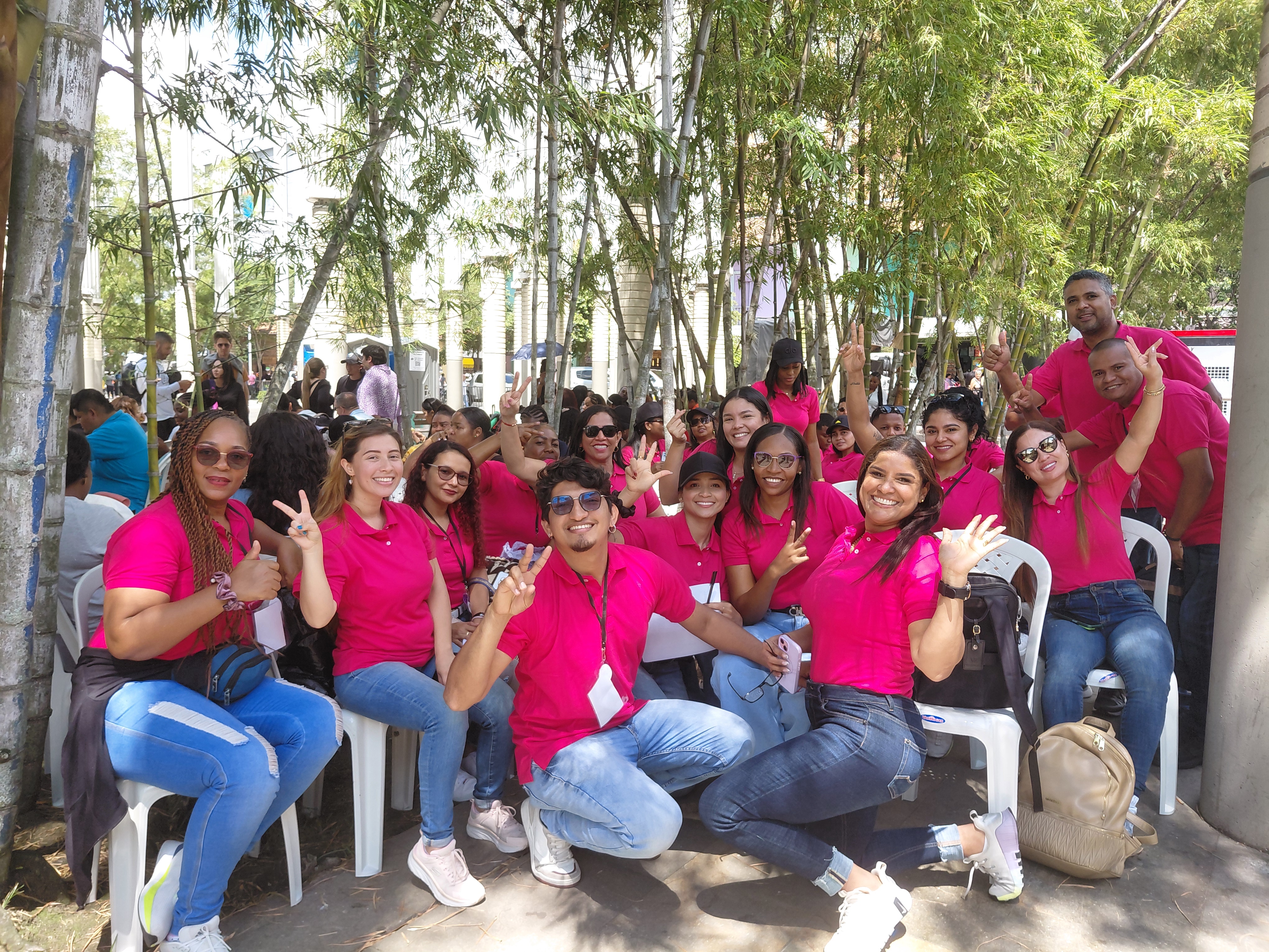 Grupo de jovenes alegres vestidos con camisetas magenta sentandos en la plaza de las luces  hacen con sus manos la señal de la victoria