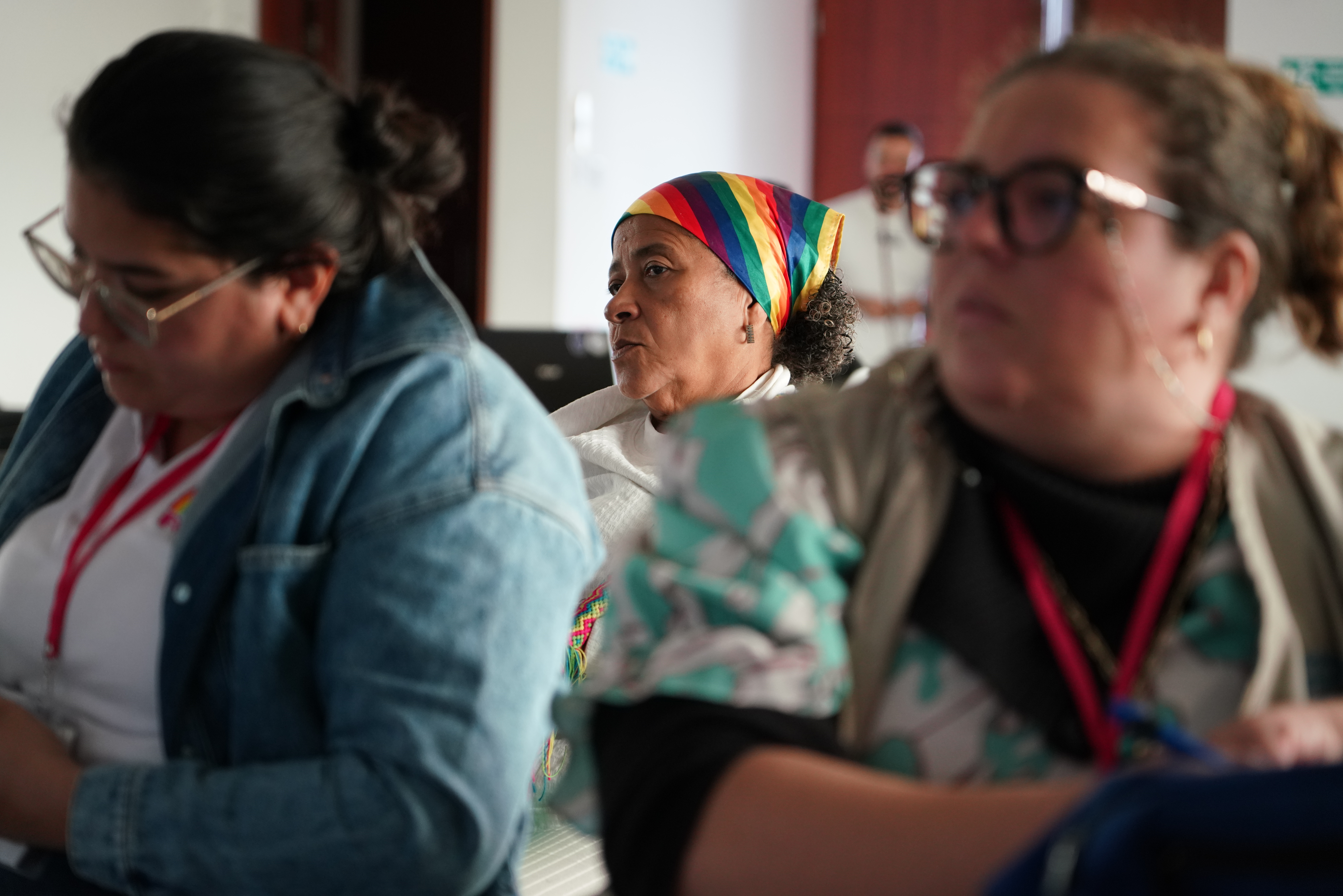 Primer plano de mujeres asistentes al taller. Al fondo en el medio el rostro de una mujer que luce una pañoleta en su cabeza con los colores LGBTIQ+