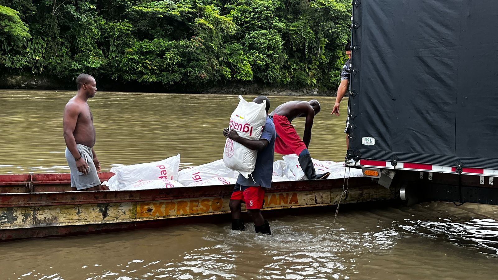 Hombres afro en pantaloneta, sin camisa, descargan un camión con las bultos de comida de MinIgualdad para llevarlas en la orilla del rio Atrato en el Chocó a una lancha artesanal. 