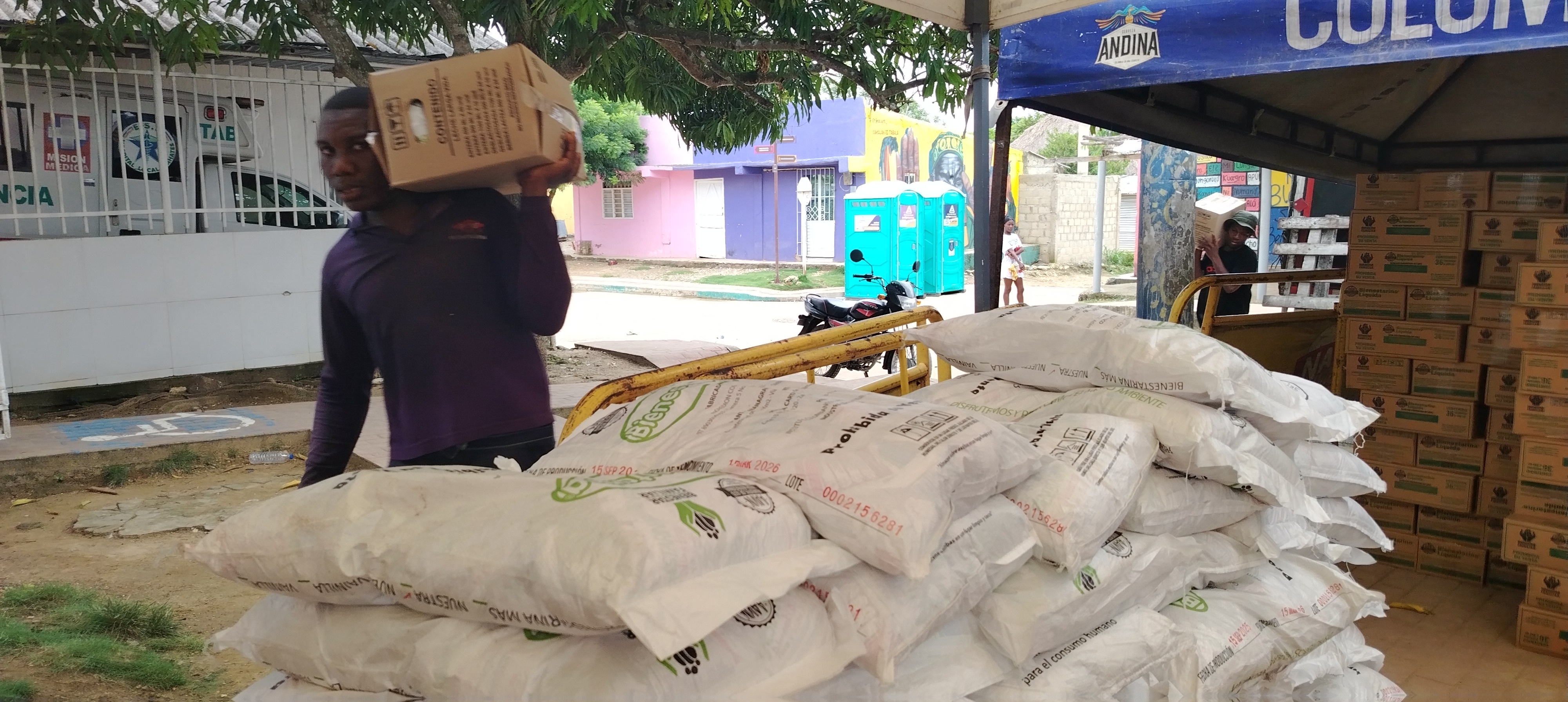 Primer plano de arrume de bultos de bienestaria. Al fondo joven afro carga al hombrio una caja de 18 litros de leche en la parte trasera de la Plaza de la Libertad de San Basiliio de Palenque