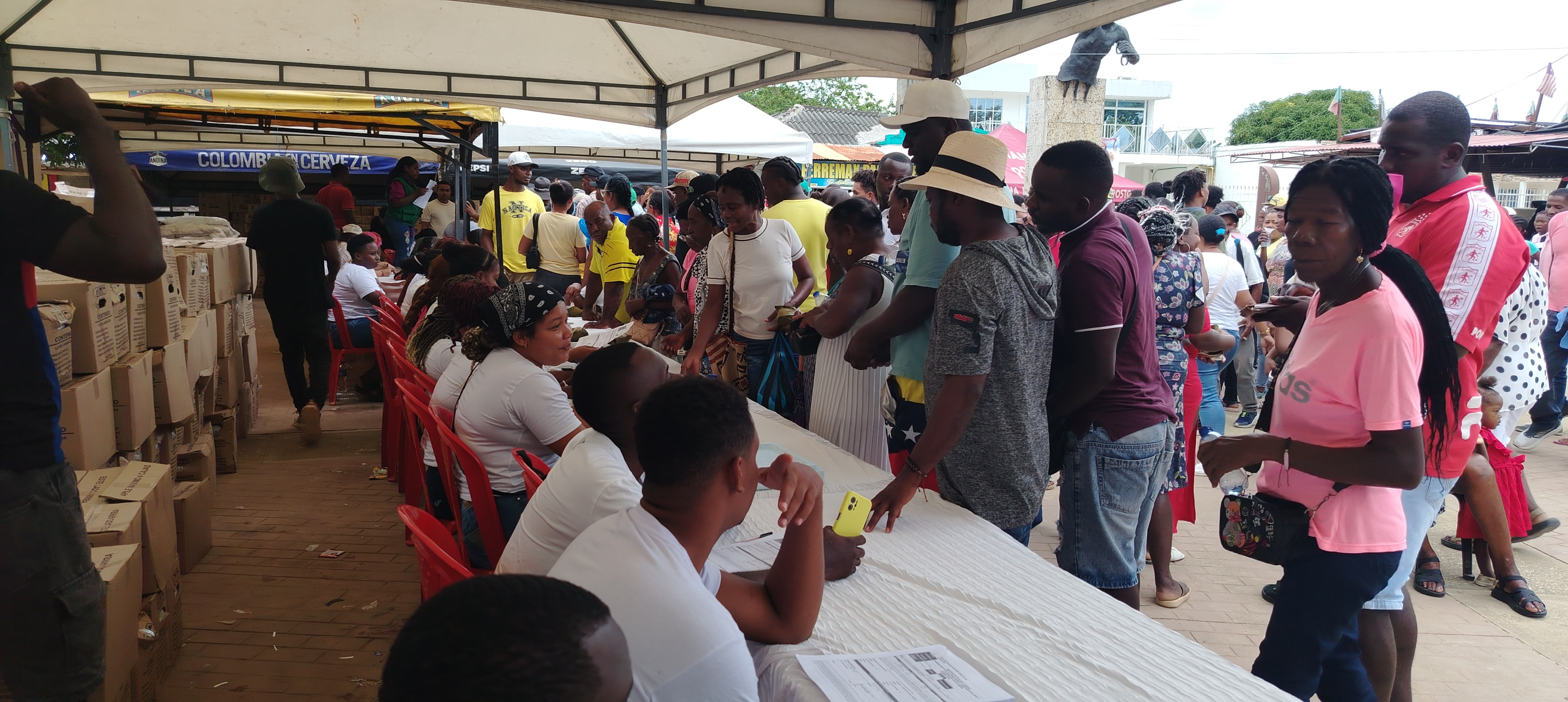 Plano general de mesas largas con funcionarios llenando planillas de registro. Del otro lado de las mesas, hombres y mujeres esperan la entrega de los productos