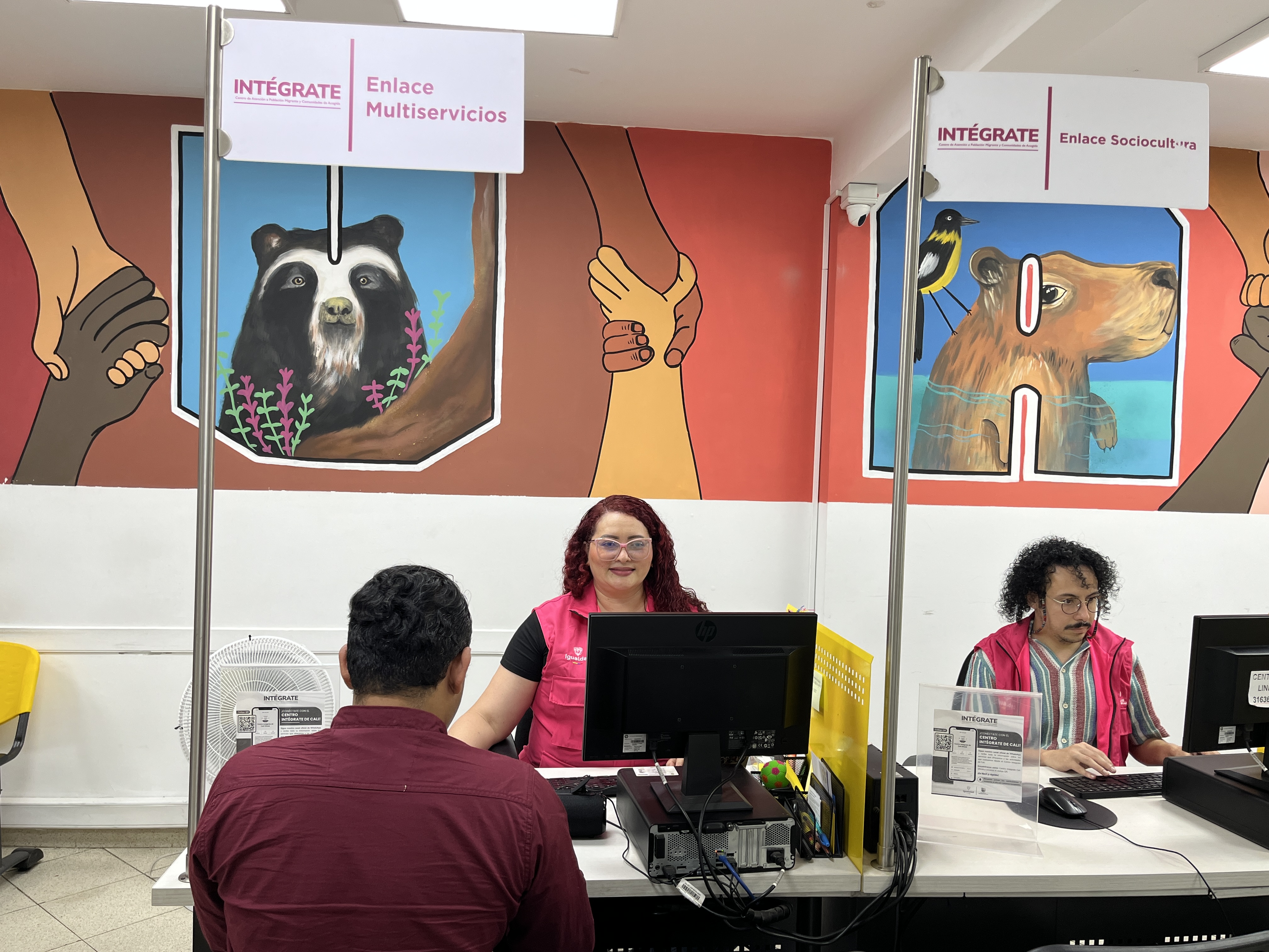 Plano medio de una de las salas de atención al público en Centro INTÉGRATE de Riohacha, dónde se aprecian dops funcionarios en sus cubículos atendiendo a personas migrantes