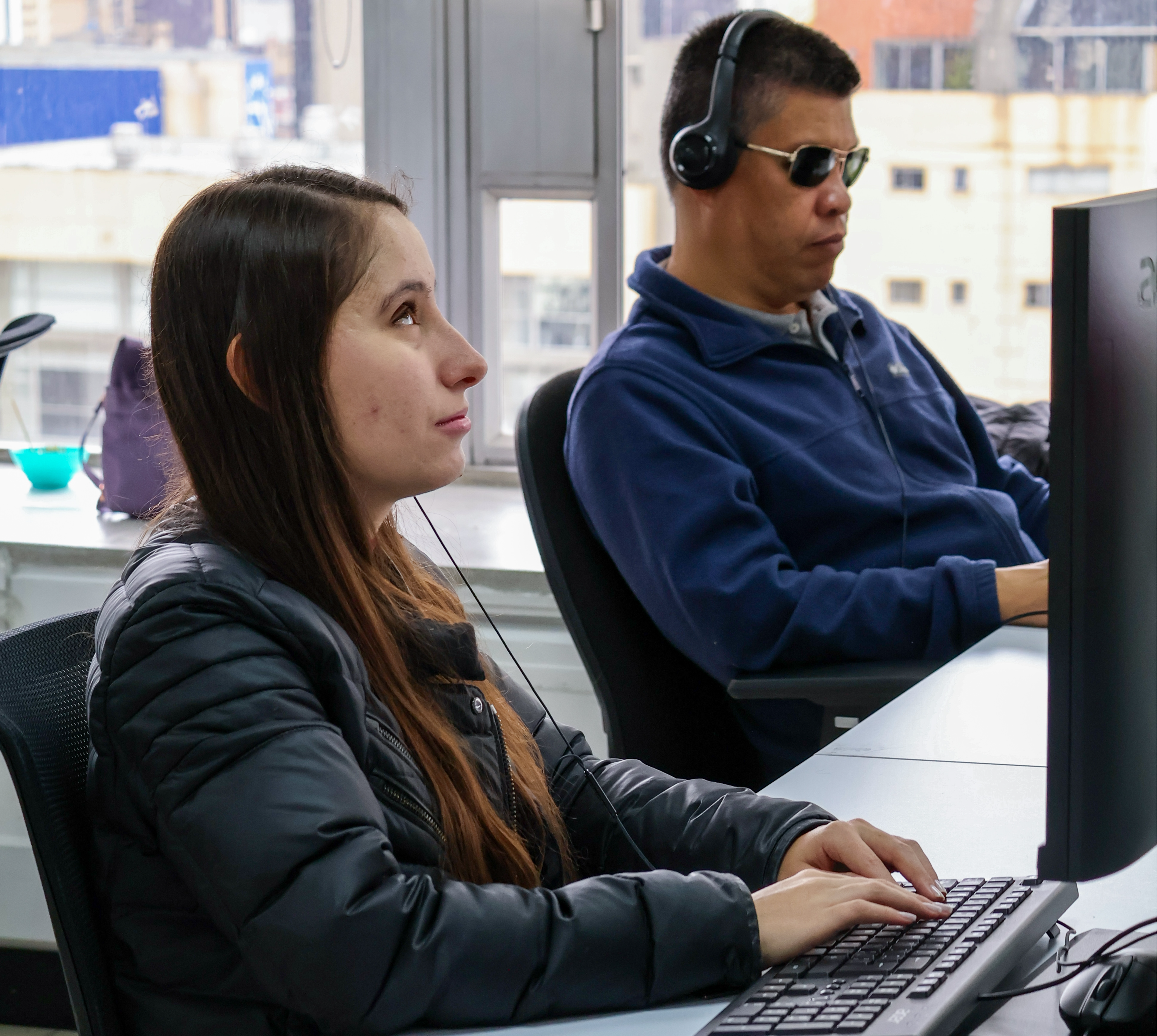 Una pareja de personas ciegas trabaja en computador en medio de un ambiente laboral
