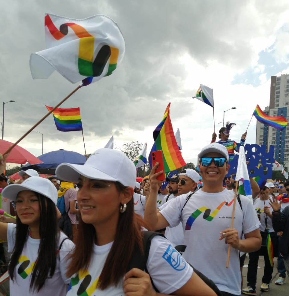 Varias mujeres y hombres jóvenes participan en al marcha del orgullo lgbtiq+ en Bogotá. Se aprecian varias banderas y gente alegre caminando por calles de la coidad