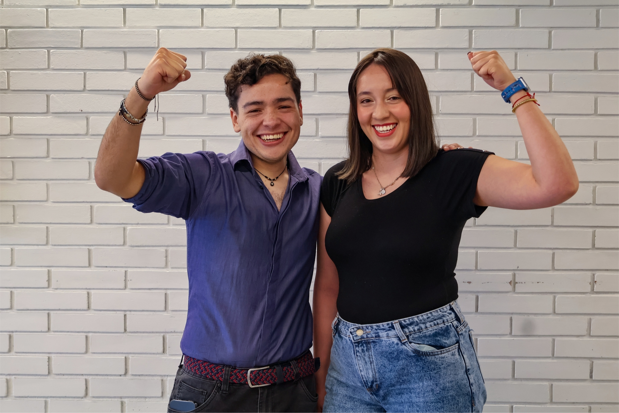 Una pareja de jovenes posa de pie delante de un muro de ladrillos blanco y cada uno levanta su brazo con el puño cerrado