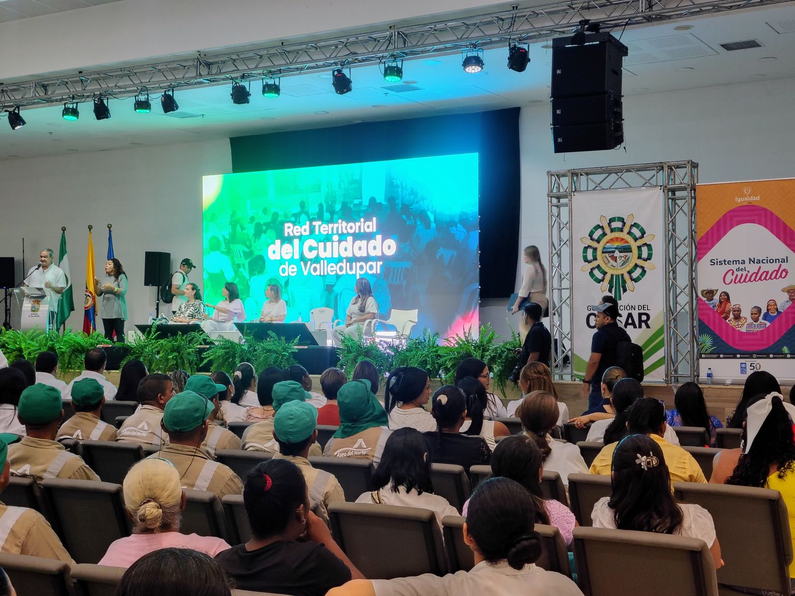 Decenas de personas sentadas en auditorio de Valledupar. Al fondo se aprecia la tarima donde personas cuiadoras están recibiendo su diploma en formación política  de la Primera Escuela de la Red del Cuidado
